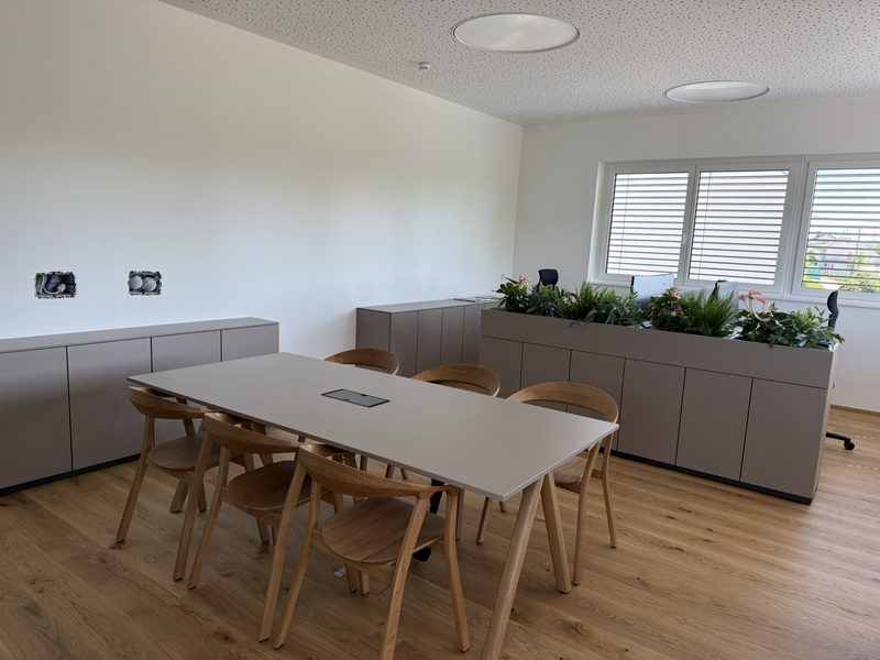 Heller Besprechungsraum mit langem Tisch, Holzstühlen, Sideboards und Fensterband mit Jalousien