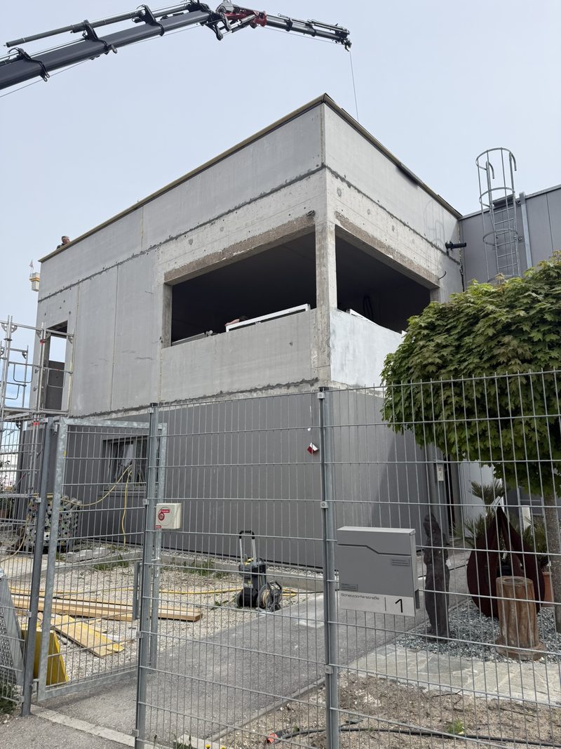 Sichtbetonkubus im Rohbau mit tiefer Fensterlaibung, Baustellenzaun und Kran